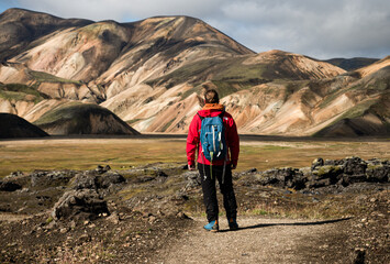 Obraz premium Adventurous man in red jacket and blue backpack hiking through stunning rainbow mountain Landmannalaugar deep inside Iceland