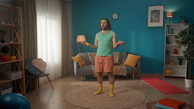 A middle-aged man with long hair. Standing and holding a jump rope. He is about to jump looking into the distance, next to him on the floor is a mat for fitness. Fitness at home.
