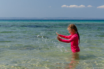 A girl standing in the sea happily playing with water. She is wearing a pink T-shirt with long sleeves.