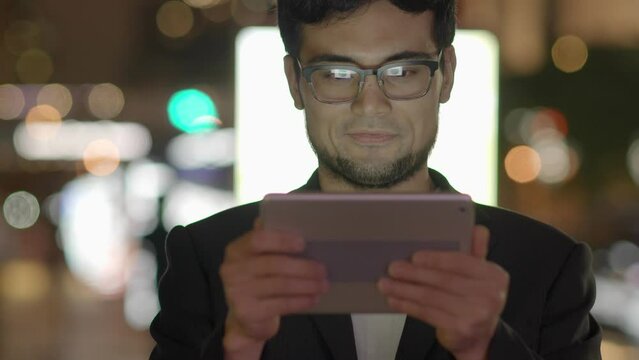 Young Asian Man With Glasses Using Tablet Screen Computer in the City at Night