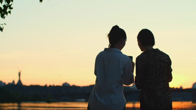 Two women take photos of river with mobile phone. Beautiful girls taking video of sunset on smartphone. Female couple of friends relaxing on the beach
