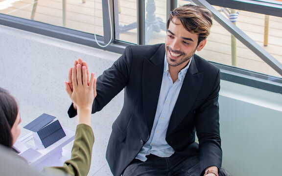 Selective focus Caucasian smart handsome businessman wearing formal suit, smiling with happiness and success with his green project with solar rooftop, giving high five, sitting in indoor office.