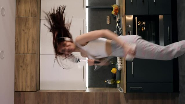 Vertical video of pretty young woman in headphones dancing in kitchen and listening music indoors Energetic dancing of a happy girl Beautiful girl enjoying music