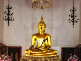 A golden statue of Buddha in a temple. The statue is in the center of the image and is seated in the lotus position. There are pink flowers in vases on either side of the statue.