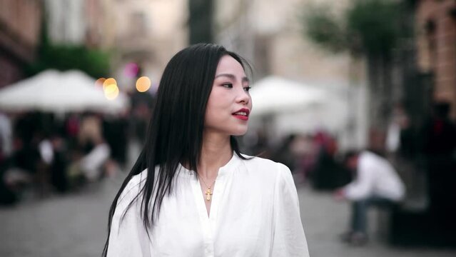 Charming young asian woman with long dark hair walking down the street and looking around at evening outdoors Happy relaxed lady walking on the city centre enjoying beautiful day alone