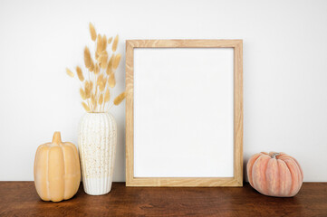 Mock up portrait wooden frame with fall grasses and pumpkin decor on a wood shelf against a white...