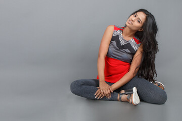 Studio shot of a young, beautiful Indian female model in casual wear wearing red and grey designer top and blue jeans against grey background. Female model. Fashion Portrait.