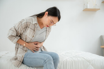  Asian woman with abdominal pain sitting in bed at home She was suffering from severe abdominal pain. Stomach discomfort and food poisoning diarrhea - obrazy, fototapety, plakaty
