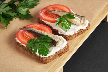 Sandwiches with sprats, tomatoes and parsley on wooden serving tray on black stone background. Appetizer, seafood, front view