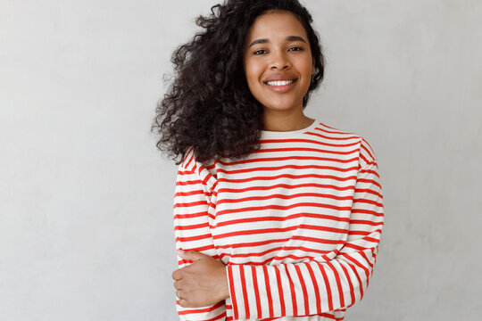 Youth problems concept. Smiling positive shy charming lovely charismatic female of 20s posing against gray studio background in striped white and red shirt, looking at camera smiling