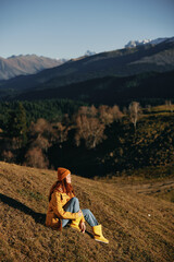 Woman full-length sitting resting on a hill smiling with teeth and looking at the mountains in a yellow raincoat and jeans happy camping trip in the spring, freedom lifestyle 