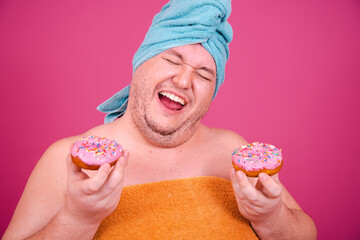 Early morning. Funny fat man gets ready for a party after a shower and eats donuts. Pink background.