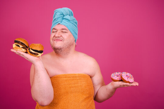 Early morning. Funny fat man gets ready to eat a burger after a shower. Pink background.
