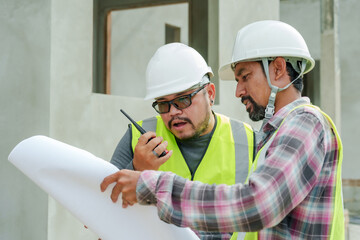 Asian construction man use walkie talkie Talk to crane operator. To control crane. engineer walks in with blueprint to give advice. At house construction site Wear a safety helmet and reflective vest.