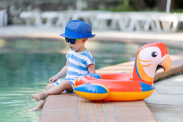 Cute kid boy wearing sun hat and sunglasses with inflatable float parrot next to the swimming pool. Summer travel vacation concept. © zphoto83