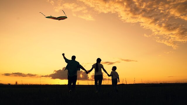 Young family is playing with kite in field. Happy family with baby in park playing with kite. Family walk on grass, child dreams of flying. People together travel, nature, silhouette. Kid wants to fly