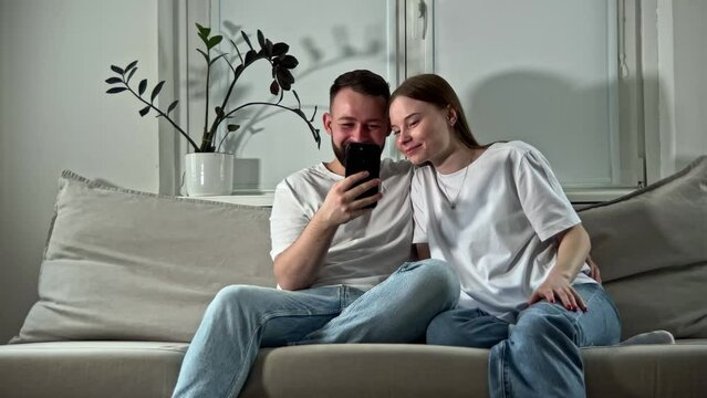Happy man and woman smiling, watching funny interesting videos and photos in online services. Young couple spending time together, scrolling smartphone and laughing