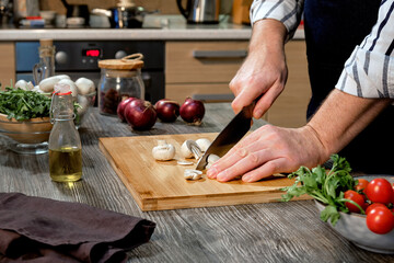 A man cuts mushrooms on a wooden board on a table with vegetables and dishes.