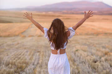 Fototapeta premium Back view of active girl raised her hands, feeling freedom in wheat field with warm sunshine.