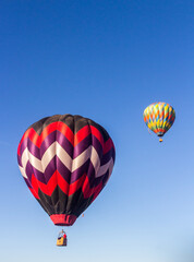 2 hot air balloons are rising in the early morning light into a blue and wispy clouded sky, 1 balloon is slightly above the balloons, The are brightly colored.