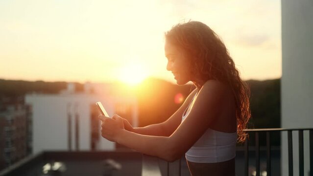 Charming young woman hold smartphone browsing scrolling app watching social media feed with beautiful sunset or sunrise at home Happy female texting on her phone enjoying leisure time at cozy balcony