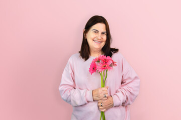 Mature Brazilian woman smiling while holding a bouquet of pink gerbera daisies, isolated against a pink background. Celebrating the life, beauty and grace of older women.