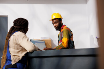 Postal service employees checking parcels invoice list on computer while chatting at checkout. African american man and woman storehouse coworkers supervising inventory management