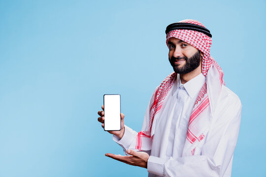 Smiling muslim man in traditional thobe presenting smartphone with empty white screen portrait. Confident arab showcasing mobile phone blank touchscreen and looking at camera