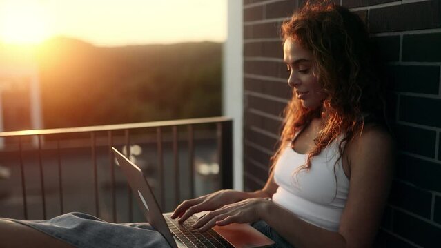 Pretty young woman freelancer working online typing browsing on laptop with beautiful sunset or sunrise at balcony Positive female enjoying great working day at cozy home