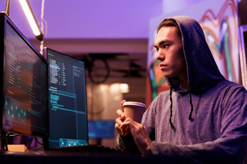 Asian hacker drinking coffee and doing ddos attack on website. Criminal breaking into network system on computer screen while holding beverage to go in abandoned warehouse