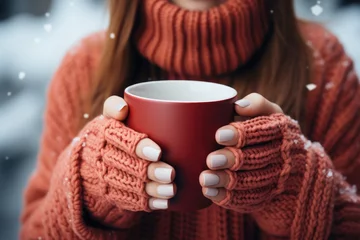  a red mug in the hands of those dressed in knitted mittens against the backdrop of a blurred snow landscape © Enigma