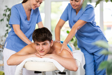 Pleased young man lying on massage table while two masseuses are massaging his back in physiotherapy room