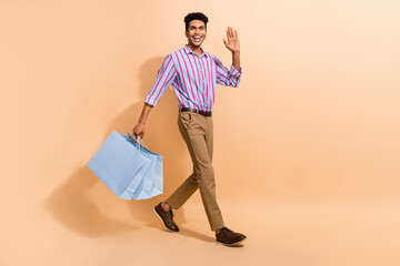 Full size photo of cool man dressed striped shirt walk shopping empty space say hi hold new clothes isolated on beige color background © deagreez