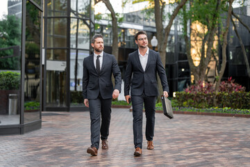 Business people discuss a project walkind on city. Two young businessmen walk outdoors city looking...
