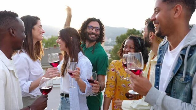 Multiracial group of friends having fun dancing and drinking red wine while celebrating on rooftop party at sunset over city background.