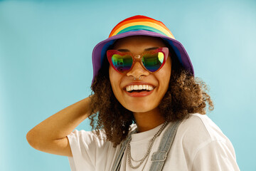 Smiling woman in rainbow cap and heart-shaped glasses looks camera over blue studio background