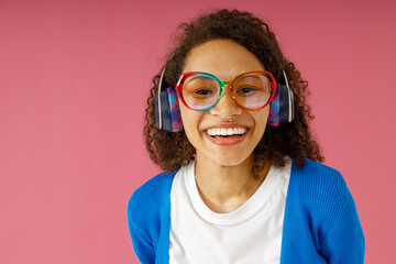 Smiling woman in headphones and glasses looking at camera while standing on pink studio background