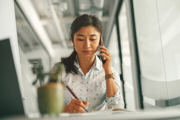 Businesswoman talking phone with client and making notes while sitting in office. High quality photo