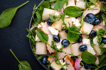 Gourmet salad with juicy sweet melon, grilled chicken, red onion, cheese, blueberries and mixed herbs, black table background, top view