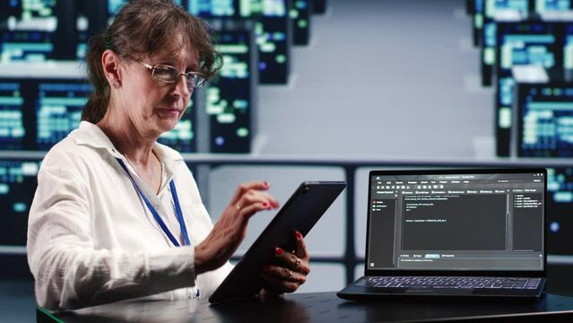 Older employee in technologically advanced server room running scripts on laptop and tablet. Tech support woman amidst data center hardware manipulates code and improves cybersecurity