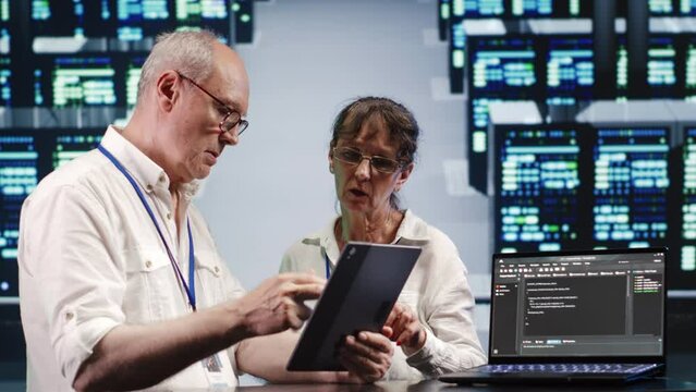Hardworking engineers in high tech facility, solving tasks on tablet while running scripts on laptop terminal. Dilligent technicians amidst blade servers, accessing databases and manipulating code