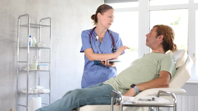 Friendly female doctor holding document folder and engaged in conversation with male patient lying on examination couch in medical office, discussing procedure details or treatment options. High
