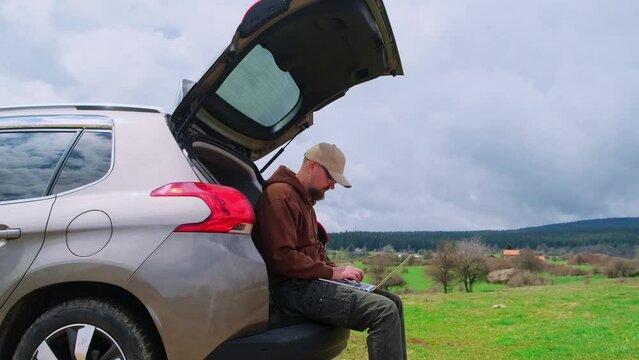 Bearded man traveler wearing cap typing on a laptop keyboard while sitting in the car trunk. Male freelancer works during road trip. Digital nomad lifestyle concept.
