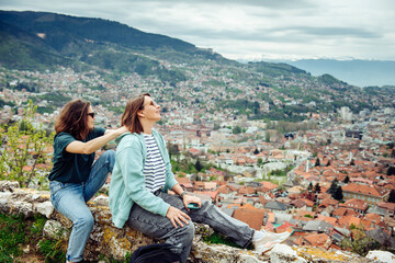 Two cheerful traveler girls in dark glasses are having fun and doing massage against the background of Sarajevo, a beautiful European city in the mountains