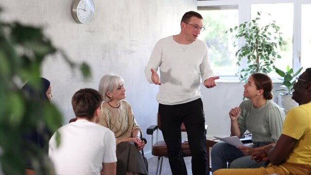 Smiling friendly man conducting informal work meeting with mixed age group of colleagues of different nationalities sitting on chairs in circle in office, discussing work-related situations. High