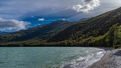 The beautiful emerald lake is surrounded by mountains. Green forest on the slopes. The waves are foaming on the pebbly shore. Clouds in the blue sky. Lago Roca. Tierra del Fuego National Park.