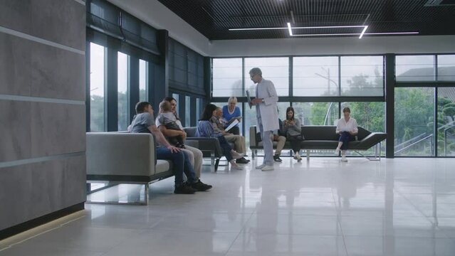 Diverse people sit on sofas in clinic lobby area, wait for doctors appointment. Doctor speaks with patients about medical examination, test results. Waiting room in modern medical center. Healthcare.