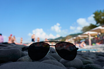 Sunglasses lie on sea pebbles. In the background are umbrellas, vacationers, mountains and a cloudy sky. Selective focus.