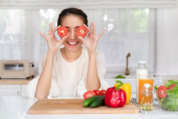 Happy young latin lady having fun and covering eyes with tomato halves, fooling around in kitchen interior, looking and smiling at camera while cooking dinner