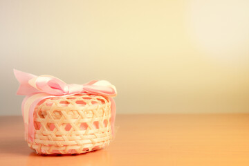 A woven wooden basket with a pink bow-tied lid placed on the left side of the warm, white background